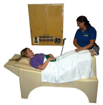 colon hdyrptherapy bed with you woman covered with a sheet. Therapist stand at her feet next to the water flusing unit hanging on the wall